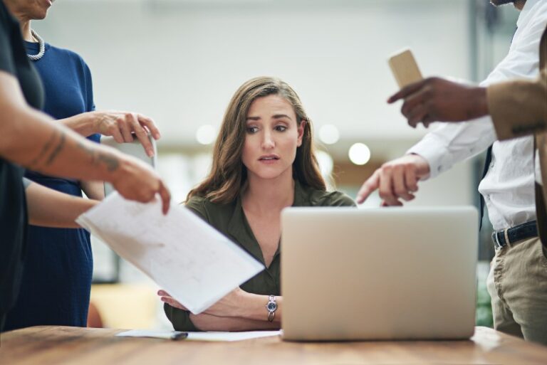 A woman sitting at a table, looking stressed, while multiple people point at her and hand her documents, symbolizing workplace pressure or discrimination.