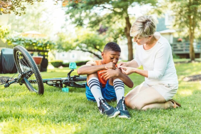 A child sits on the grass with a scraped knee next to a fallen bicycle while an adult assists him, illustrating a common area injury in an apartment complex setting.