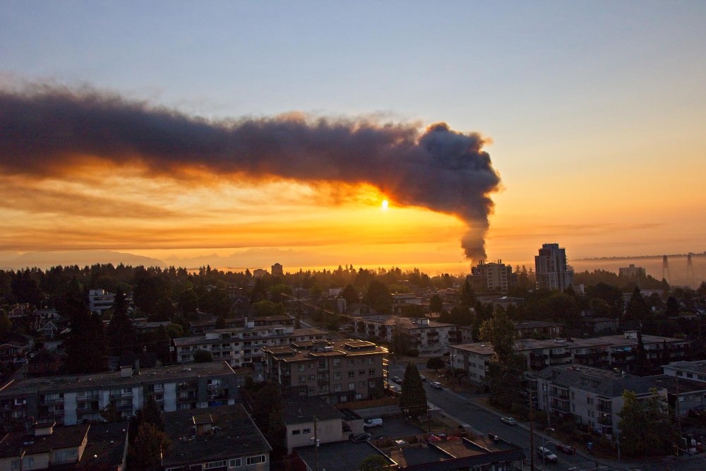 Una densa columna de humo negro se eleva al cielo durante el atardecer sobre un vecindario residencial, lo que resalta los posibles riesgos de monóxido de carbono por incendios o incidentes de combustión.